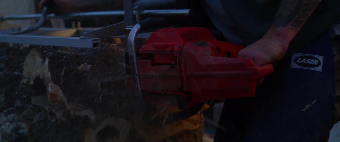 A man uses a chainsaw to cut lumber outdoors in the evening