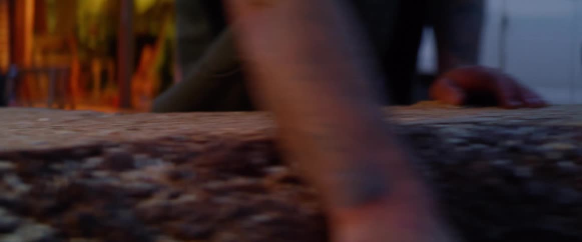 A man with a vest rubbing his hands across a piece of wood with a vehicle in the background
