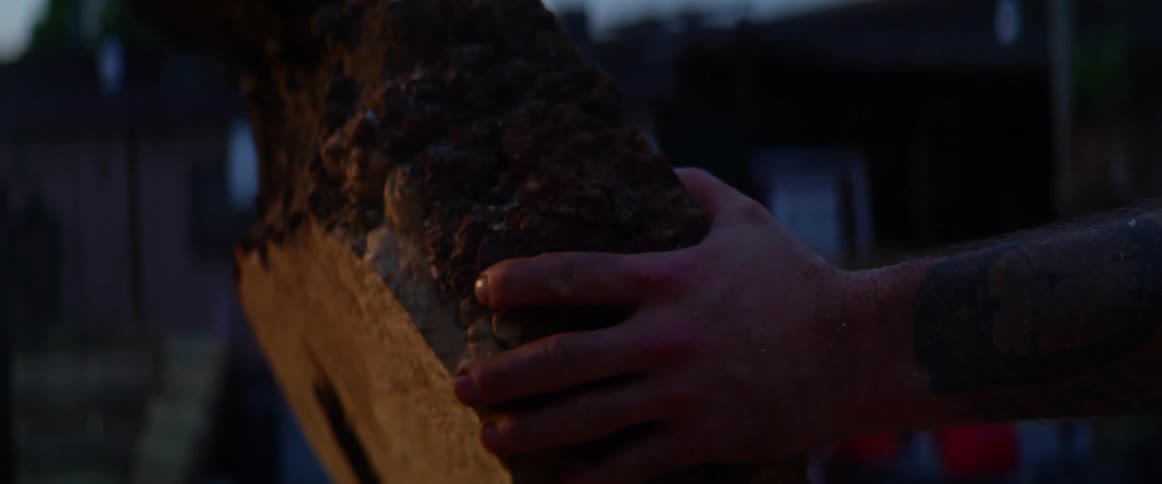 A man quickly reaches out to steady a large piece of cut wood leaning against a large wood stump in a workshop