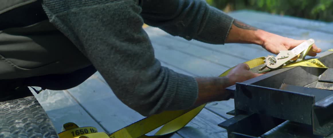 A man secures a large piece of metal to a truck bed with a ratchet strap