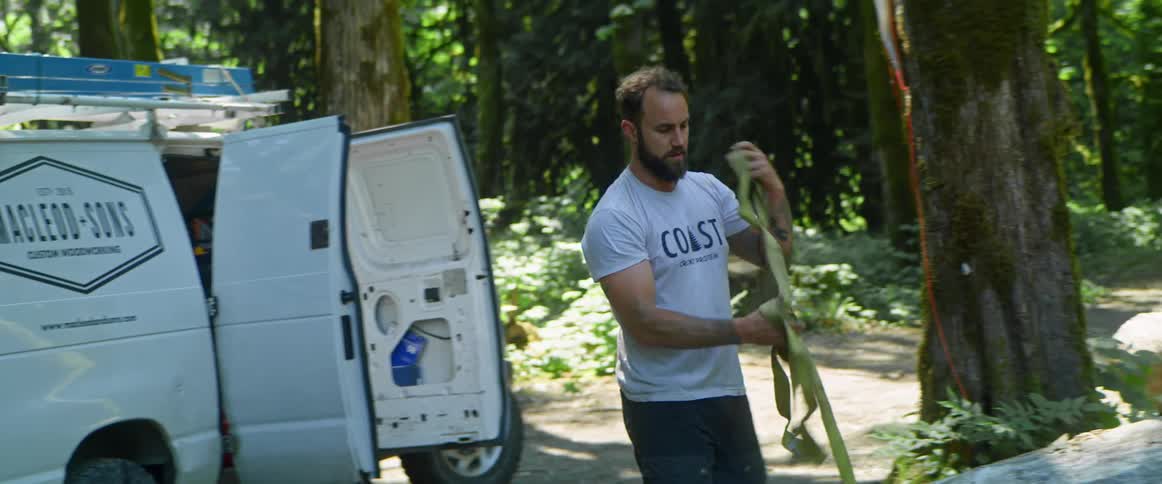 A man pulls out a large strap from his commercial van in a heavily wooded area