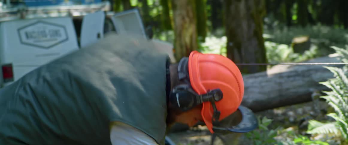 Man with an orange helmet and headphones pulls up on the starting cord of a chainsaw
