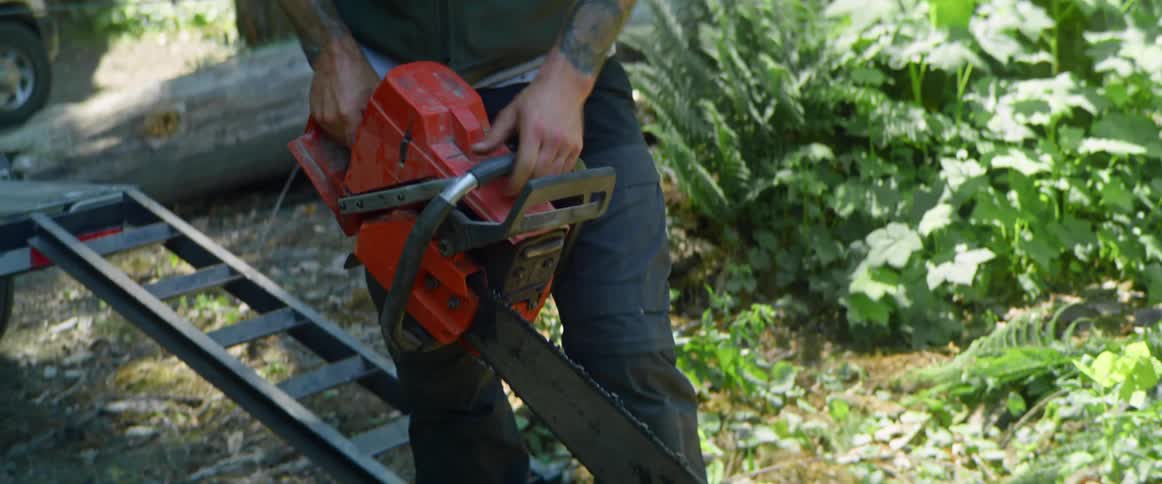 A man in a heavily wooded rural area picks up a chainsaw to cut a large fallen tree