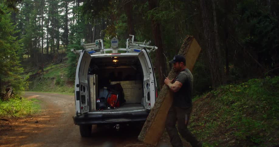 A man carries a large slab of wood to the back of his work van thats sitting on a rural gravel road