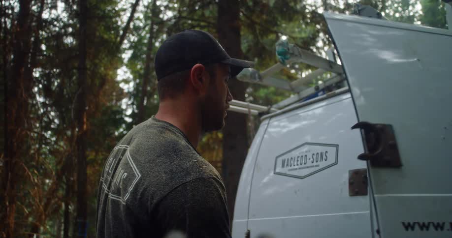 A man with a hat closing the back of a vehicle with trees clouds and the sky in the background
