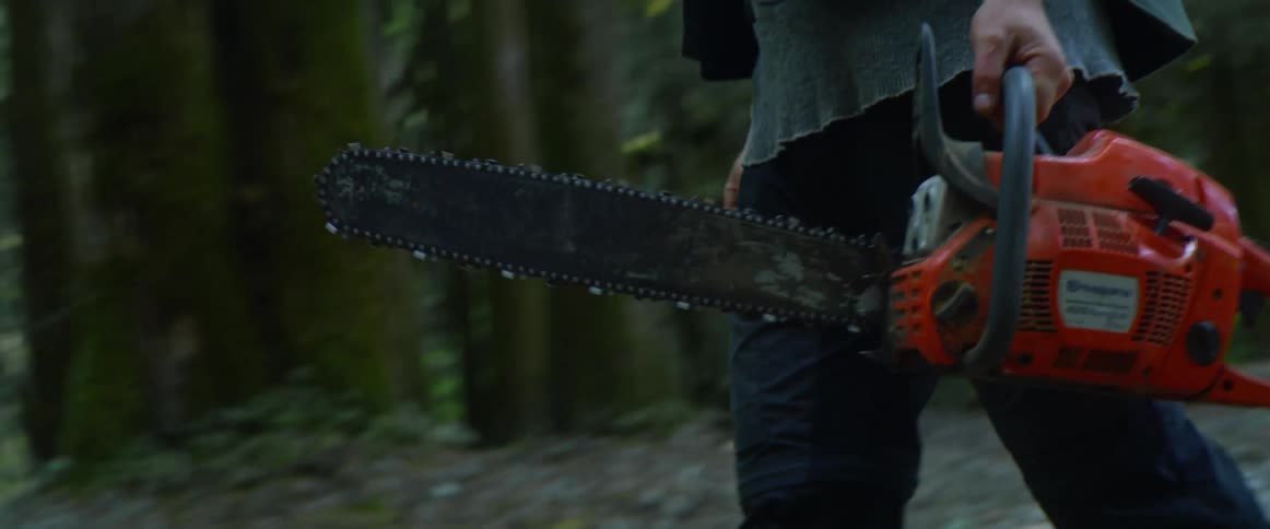 Man walks past trees in forest carrying a chainsaw