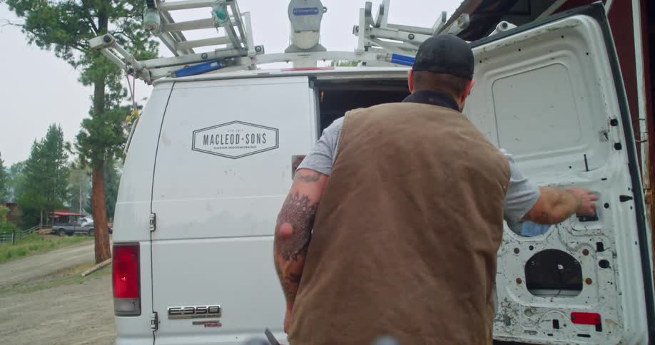 A burly man with tattoos carries a large power tool and puts it into his work van
