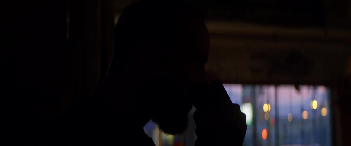Man standing in dark room drinks coffee after looking toward city lights visible through curtains