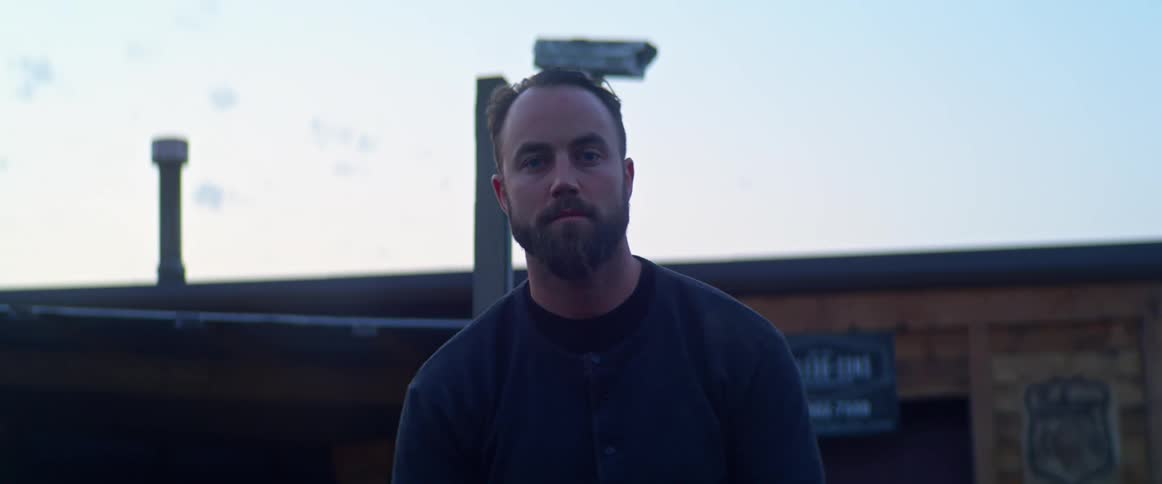 A man looks into the camera while standing outside of a wooden building during the day
