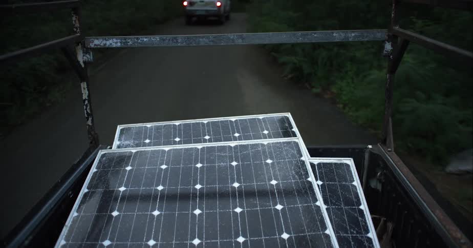 A truck with solar panels in its bed travels down a narrow road through a woodland in the evening