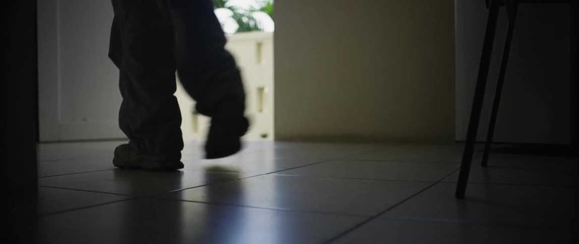 A person lifts a backpack off a tiled floor before walking through a door leading outdoors