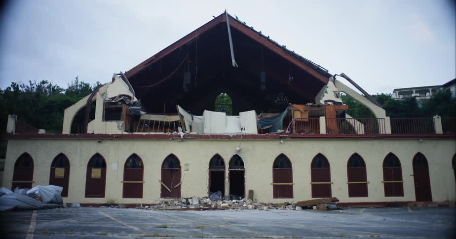 Partially destroyed building with empty parking lot is separated from another building by trees