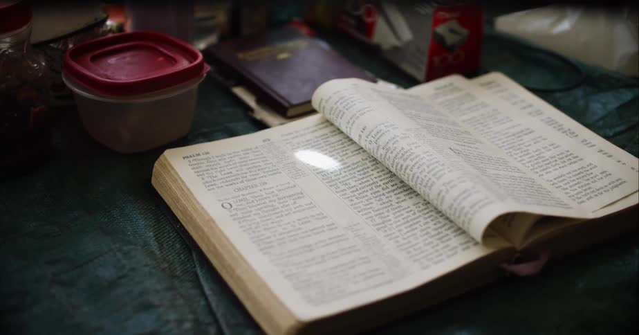 A large book lays open on a tabletop as a container and other objects sit nearby