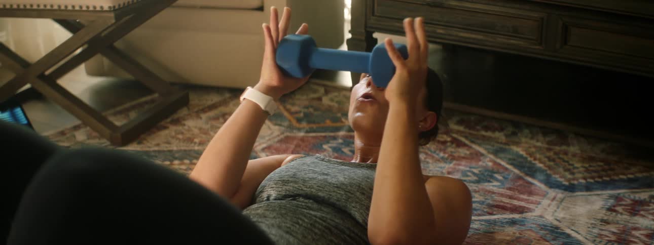 A woman holds a weight in her hands and does sit ups on the floor