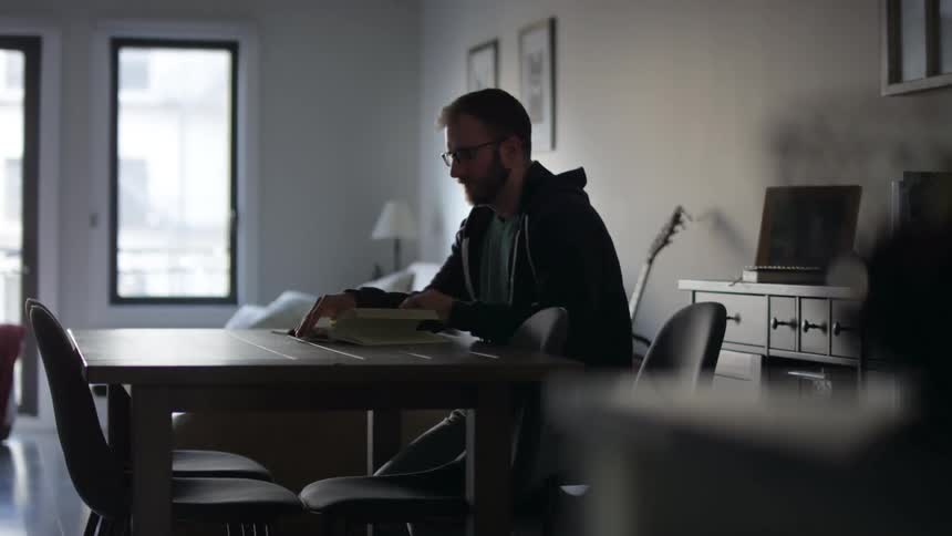 Man with a beard and glasses sits at a table and reads a book in a room
