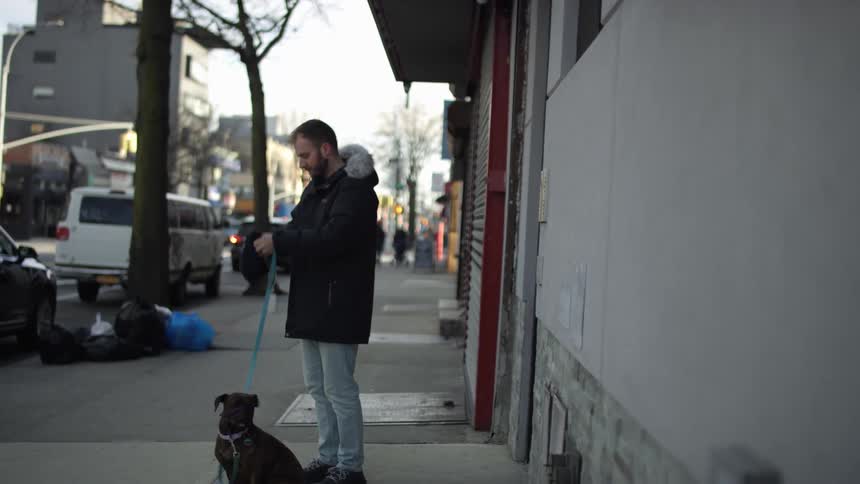 A man walks outside of a city building with his dog and puts a beanie on his head