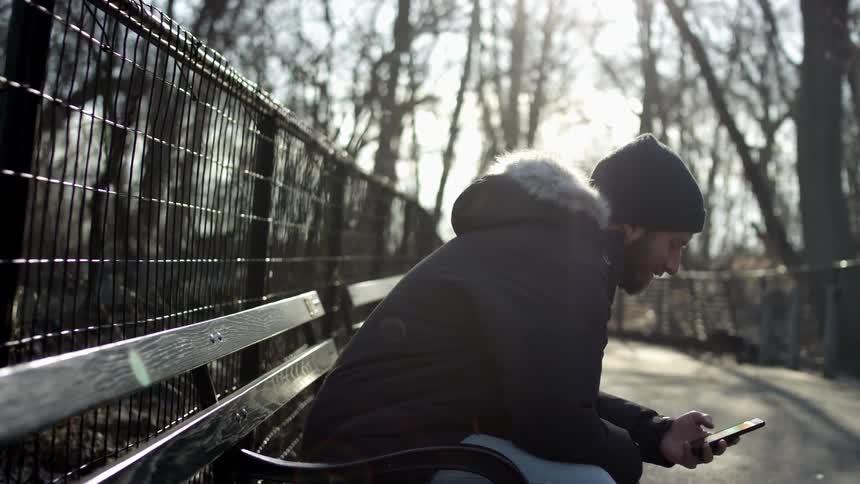 A man sitting on a bench on a walkway in a woodland throws a stick and looks at his smartphone as a dog chases after the stick
