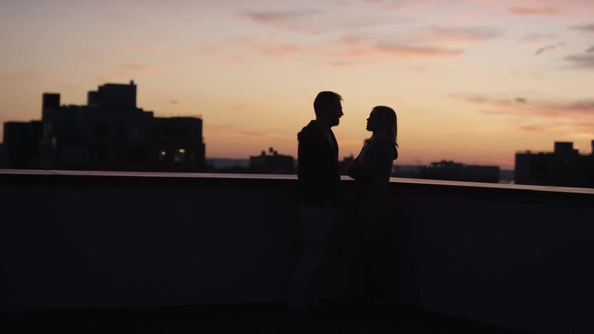 A man and woman talk while standing on the rooftop of a tall building in a metropolis at sunset
