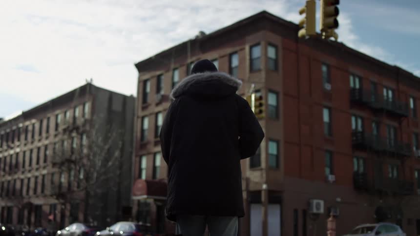 A person in a winter coat stands beneath a traffic light at an intersection in a city during the day