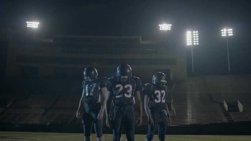 Three football players are standing on a field at night