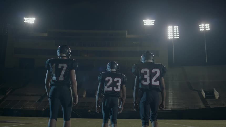 Three football players walk forward in a stadium lit up at night