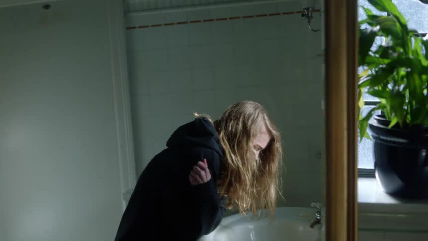 Blonde woman wearing hoodie leans over sink in bathroom