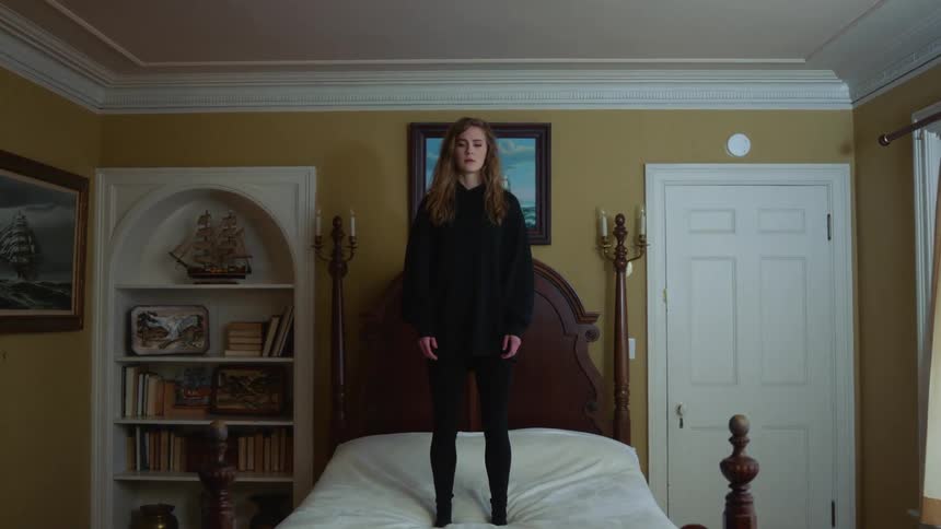 Blonde girl with a black hoodie stands on the bed in the bedroom