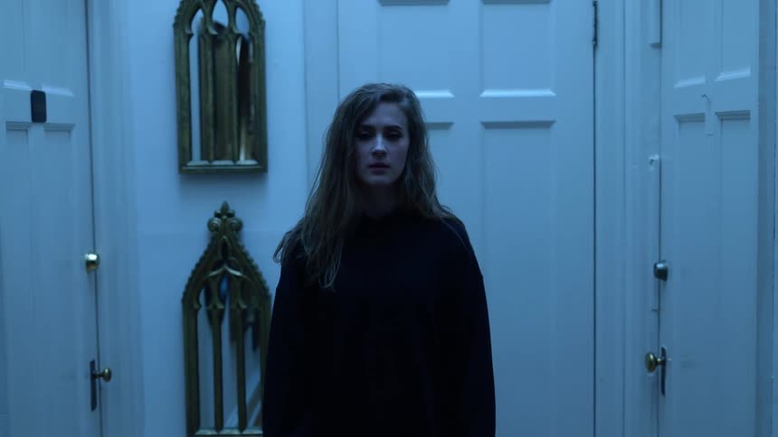 A teenage girl looking sad stands in a hallway filled with mirrors on the wall