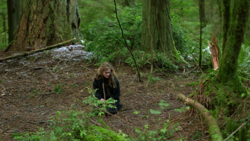 A teenage girls kneels on the ground in the forest and twists around as she acts out a scene