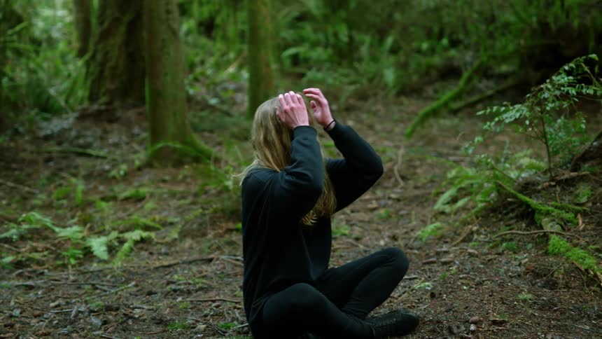 A distressed young woman sitting in the soil in a forest reaches towards the sky before clasping her forehead