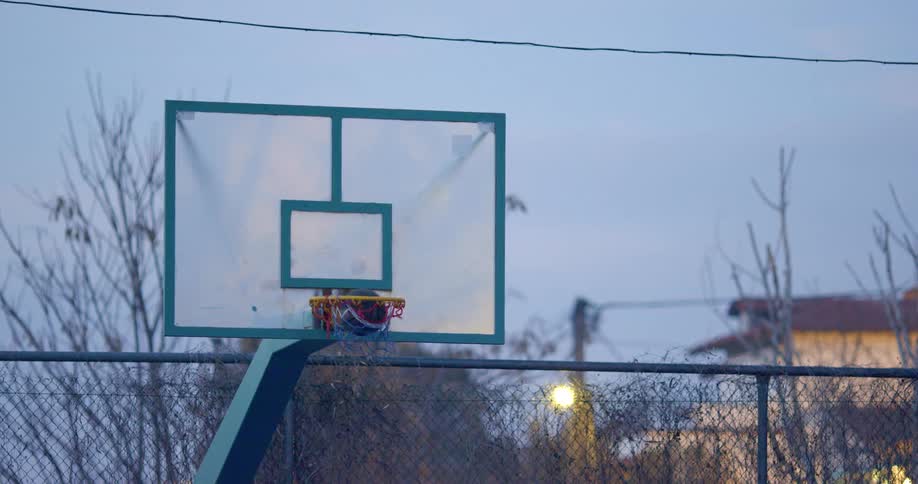 Two people throw layups while playing basketball outdoors at dusk