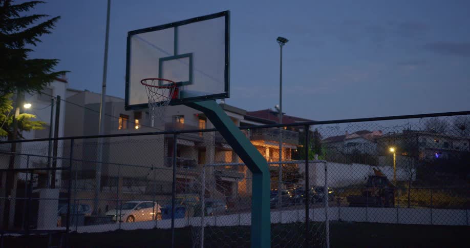 A public park with a basketball net in a neighborhood with cars parked and driving down the street