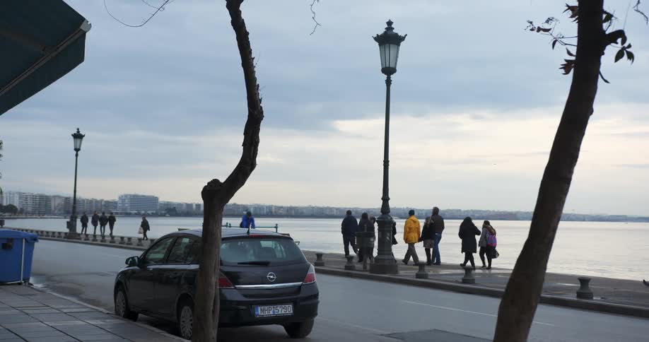 Pedestrians and traffic move along a small road bordering the ocean in a city on a cloudy day