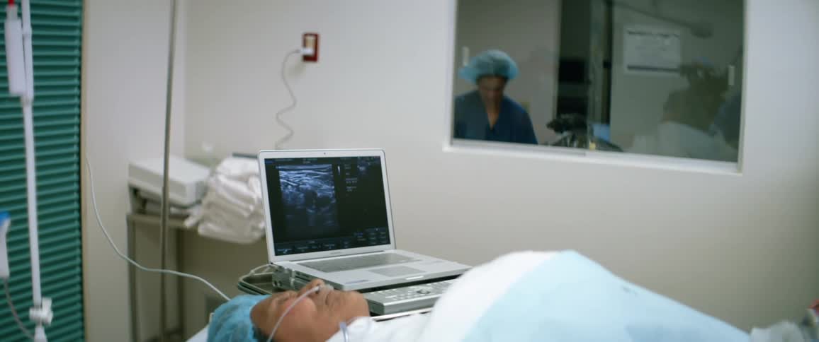 A patient lies on a bed in an operating theater as a laptop displays a medical scan