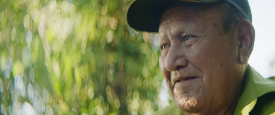 Older man wearing baseball cap stands outdoors near tree