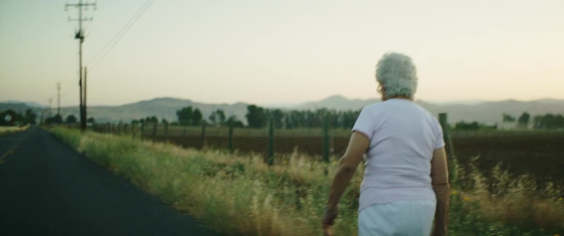 An elderly woman walks along the shoulder of a remote country road