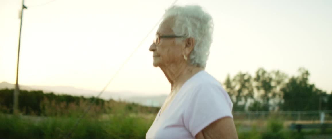 Elderly woman walks along along river near bridge with mountains on horizon