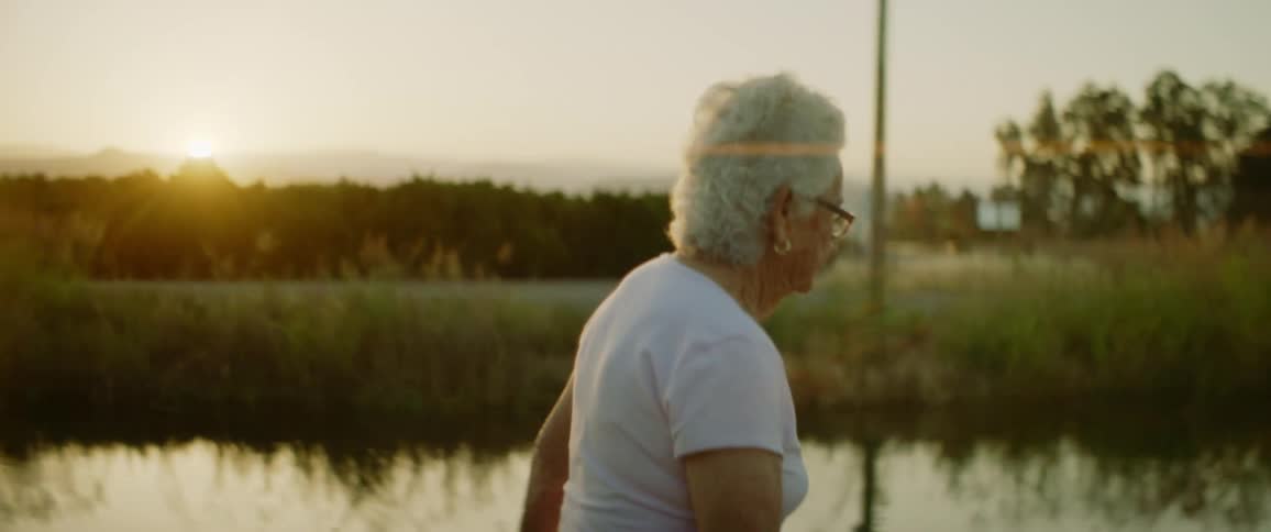 Elderly woman walks along trailnext to river while sun rises on horizon near mountains
