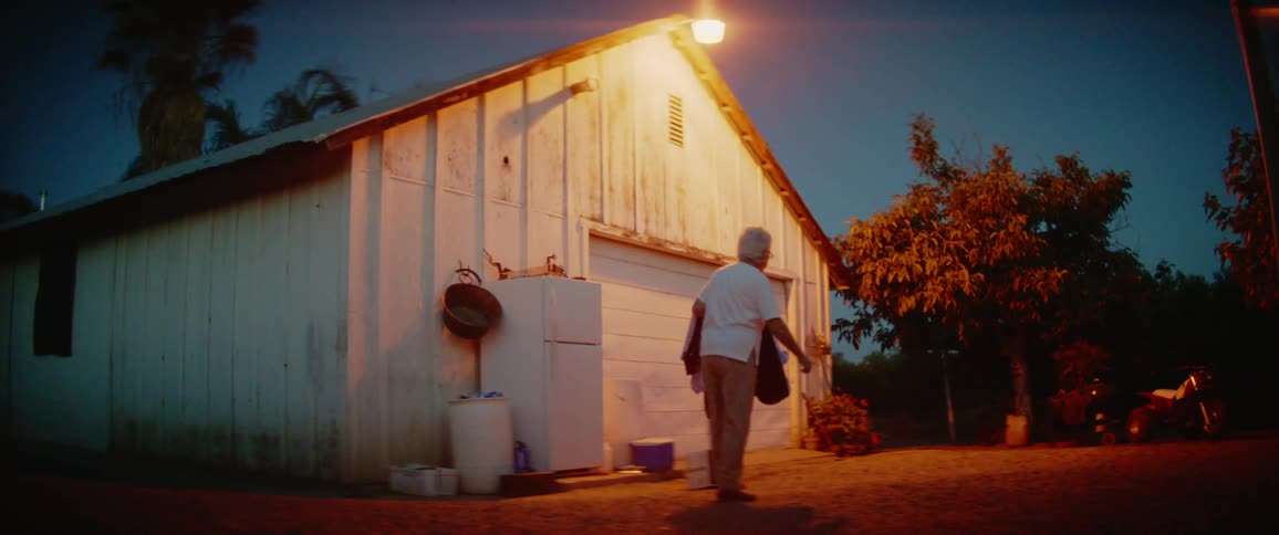 A man walks in front of a large barn at night holding a coat