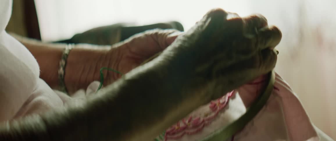 A woman sitting and putting string through a cloth with a window and curtains in the background