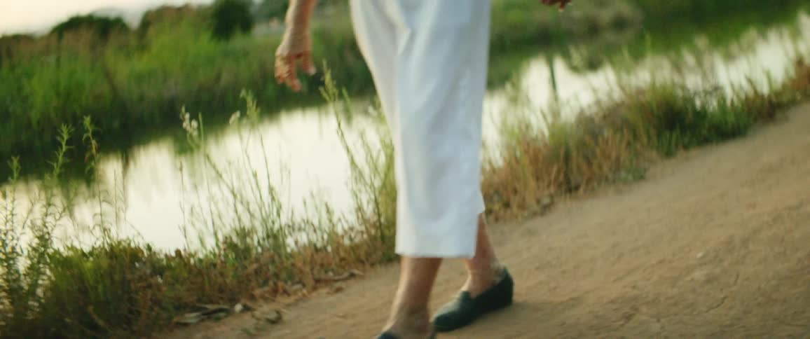 Elderly woman with white pants and a pink shirt walks on dirt road next to the river