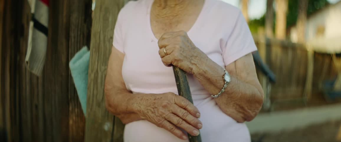 Woman holding staff leans against post in yard with wooden fence