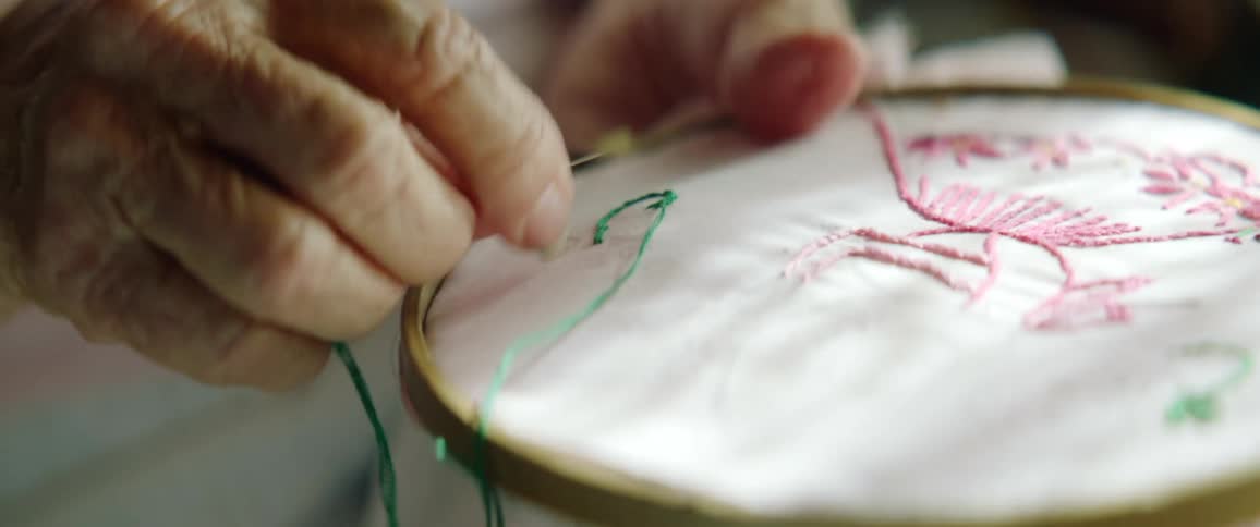 An elderly woman works on a needlepoint craft