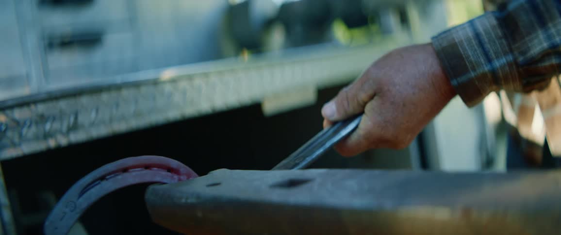 A blacksmith maneuvers a glowing hot horse shoe with a pair of tongs as he hammers it atop an anvil