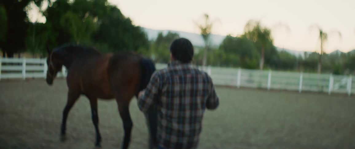 A man slowly walks after a horse as it walks around a corral