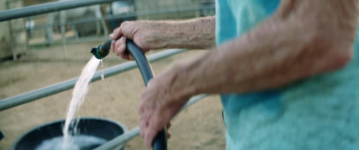 An elderly person uses a hose to fill up a bucket inside a fenced area