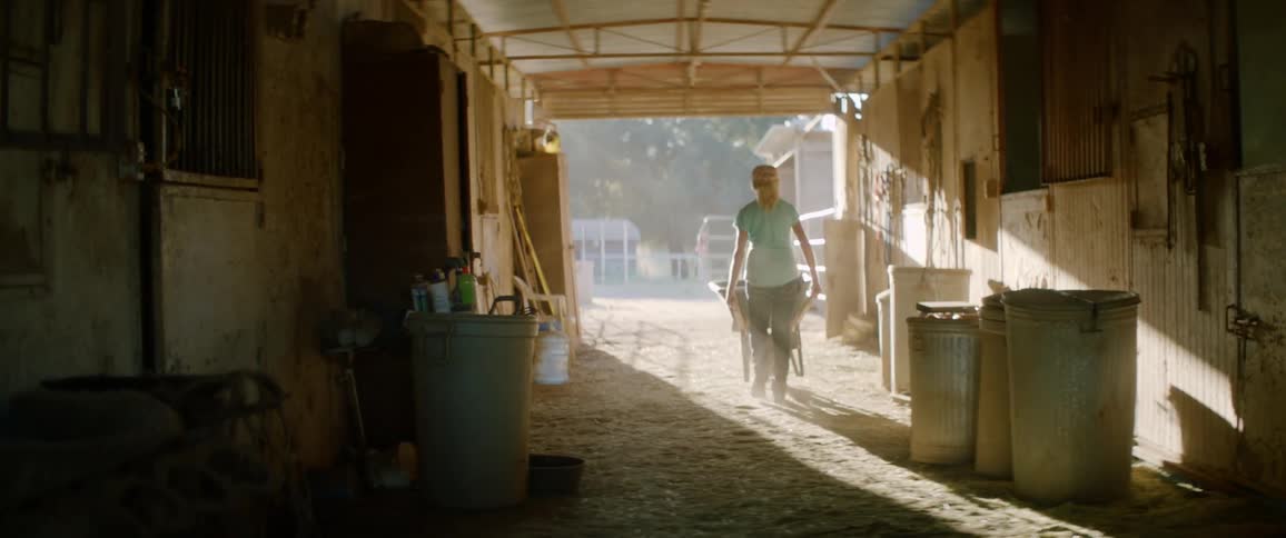 A woman rolls a wheel barrow out of a large stable barn