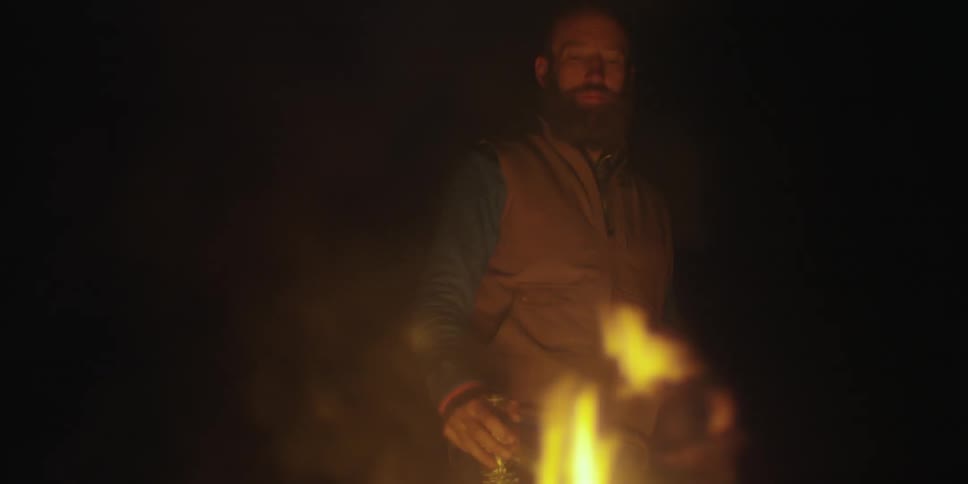 A man stands near a campfire drinking from a bottle