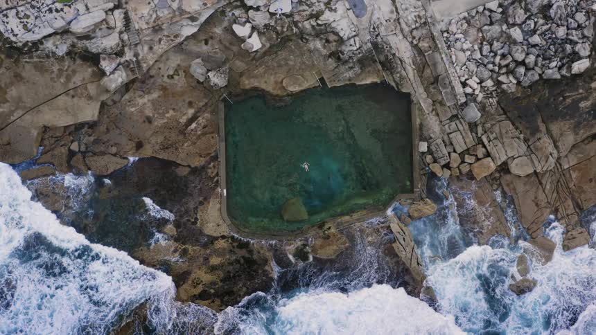 A person floats in a swimming pool on the shore as waves crash towards it