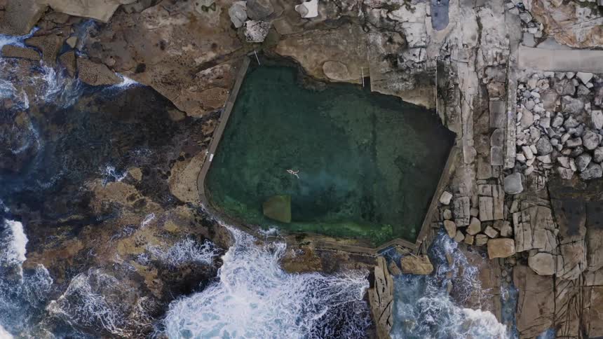 Waves crash into irregular shaped pool person is swimming in near rubble and rocks